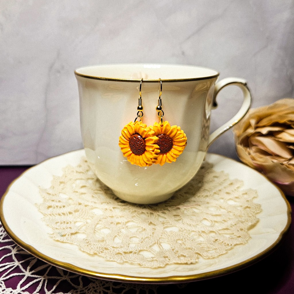 Sunflower-themed dangle earrings made from polymer clay, displayed on a teacup and saucer