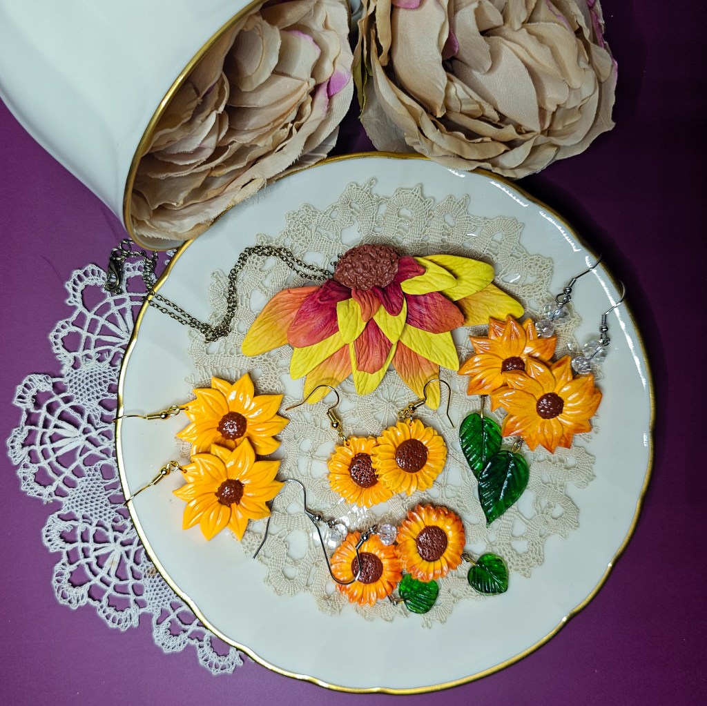 Flat lay of handmade polymer clay jewelry shaped like sunflowers, with yellow petals and brown centers on a tea saucer