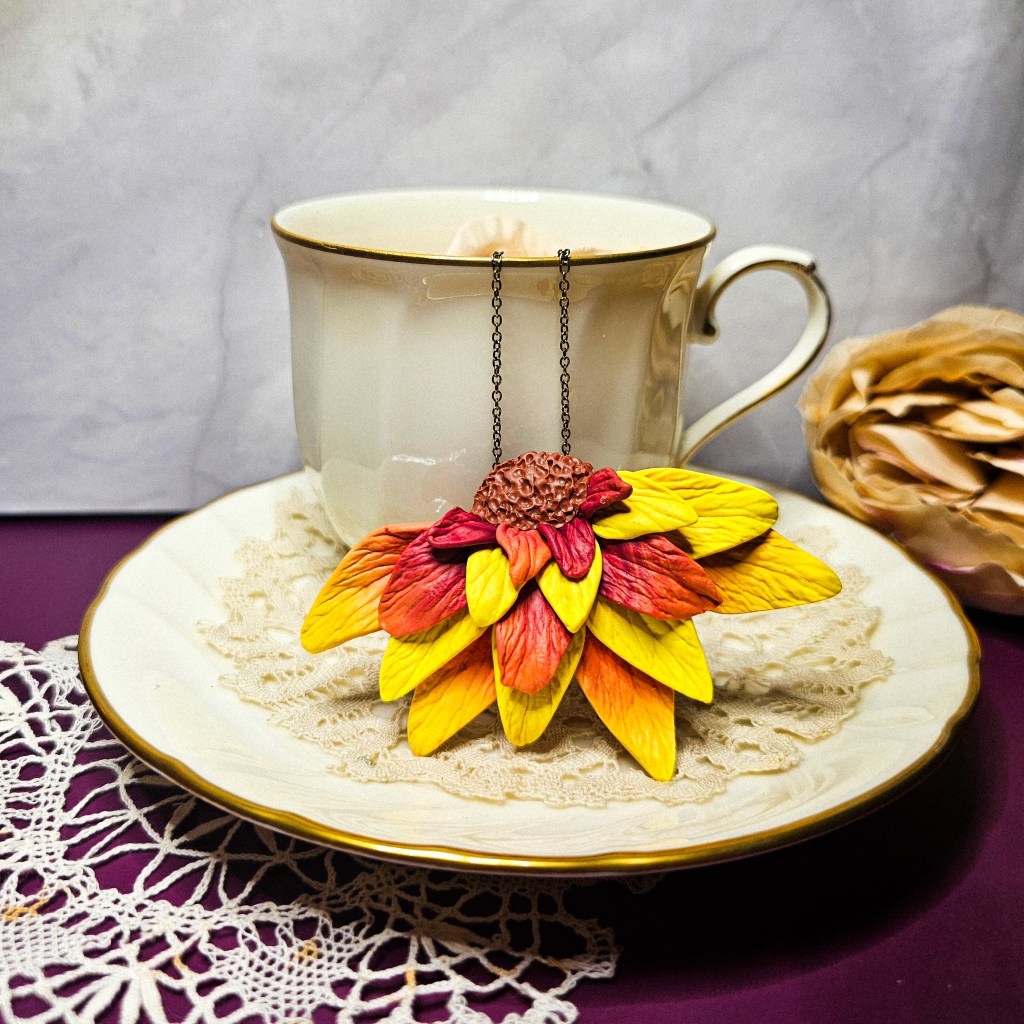 Handmade half sunflower pendant made from polymer clay displayed on a tea cup and saucer.