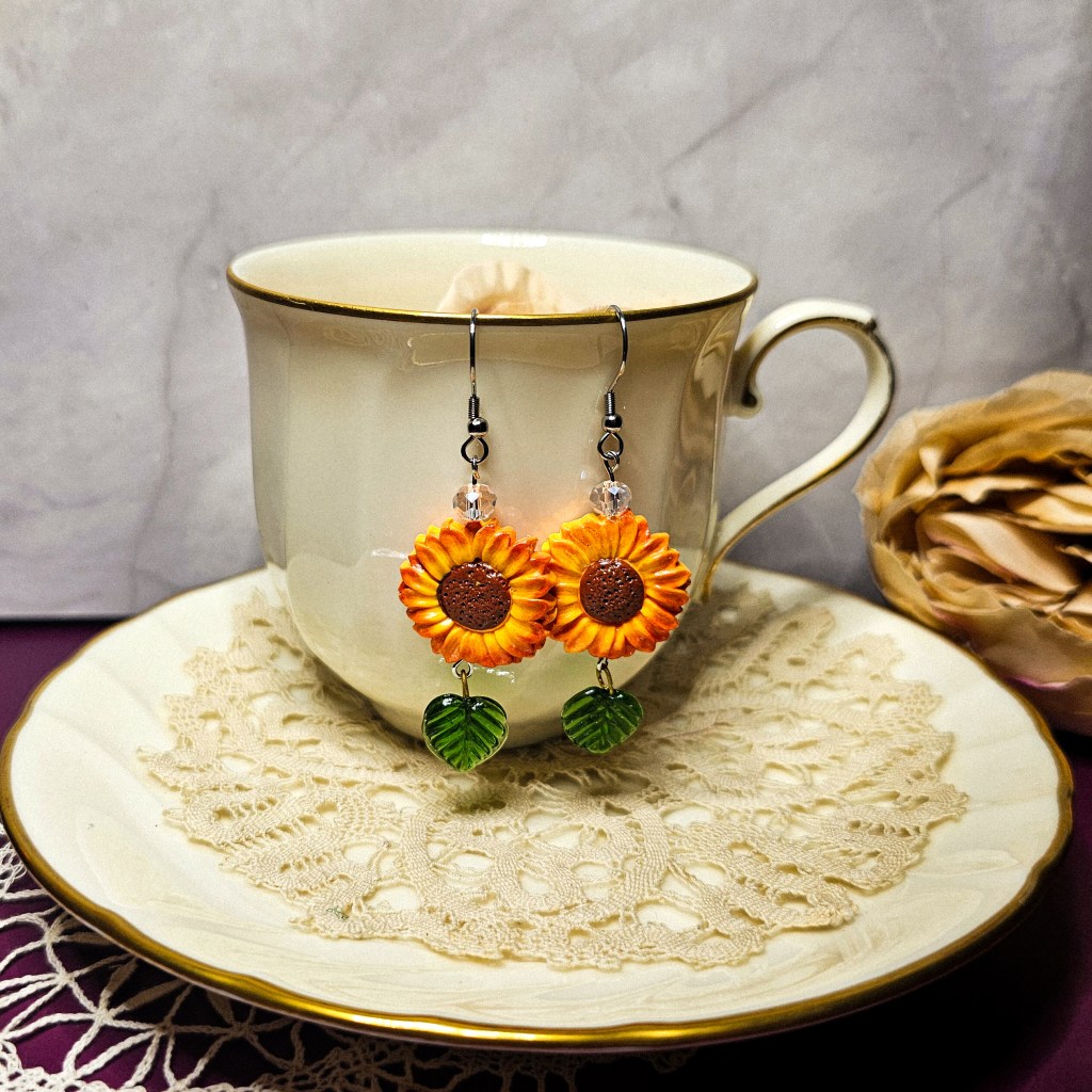Sunflower-themed dangle earrings made from polymer clay, displayed on a teacup and saucer